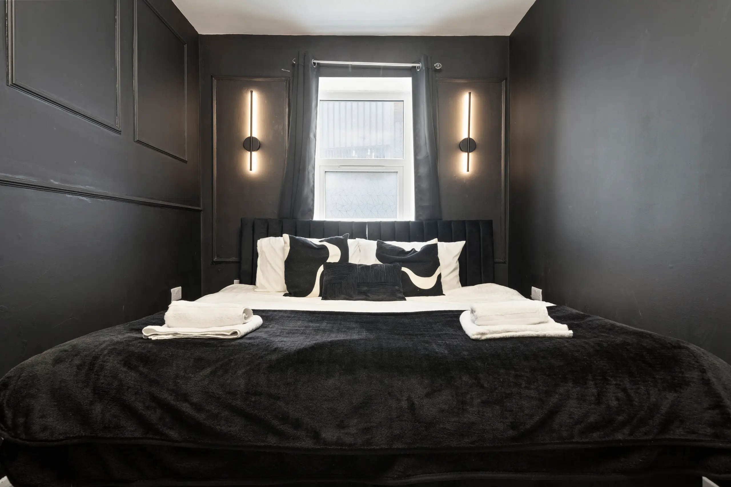 Bedroom with black panelled walls, slim wall lights, patterned cushions and folded towels on the bed