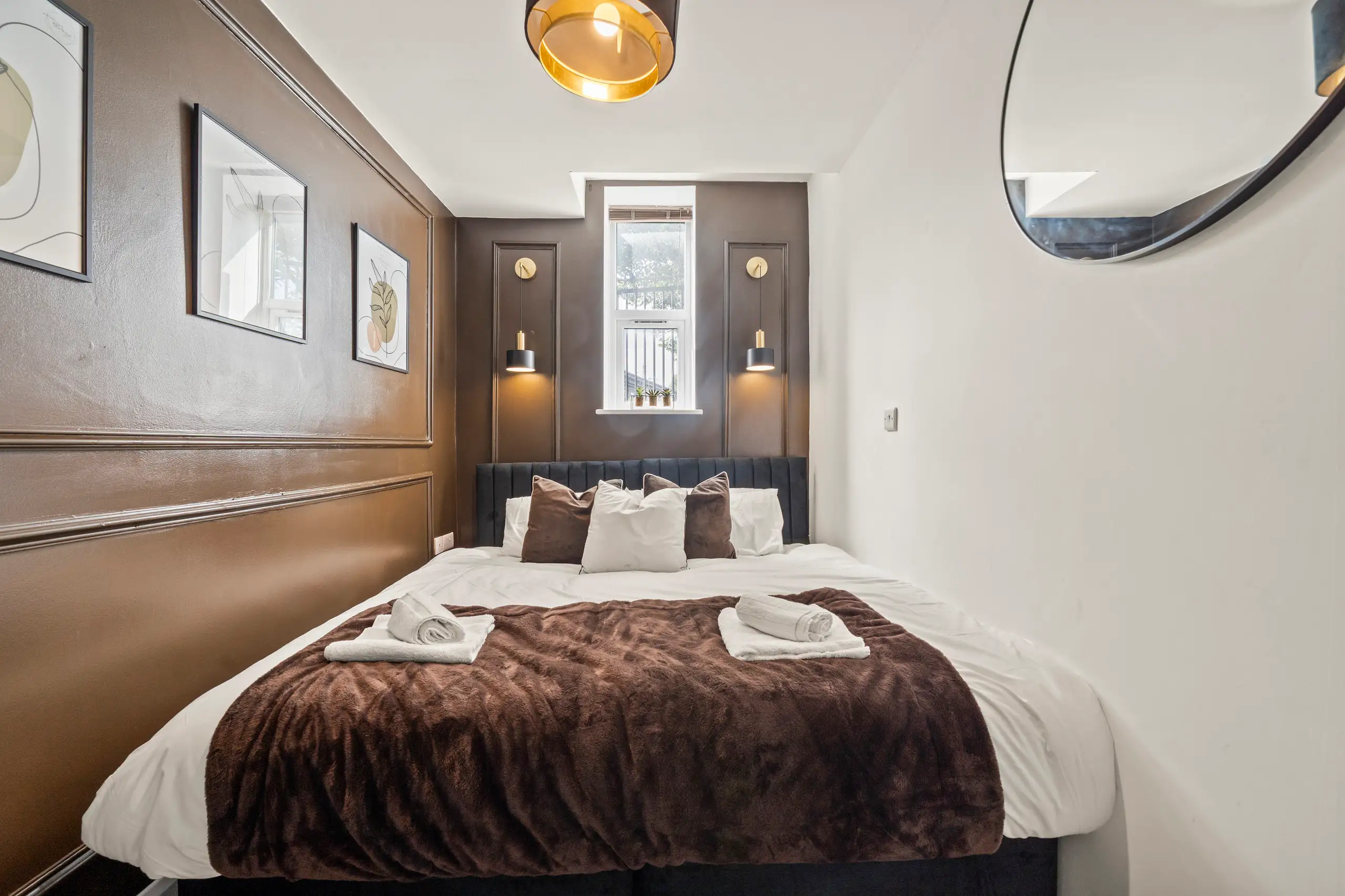 Double bedroom with dark brown panelled walls, wall lights either side of the window and folded towels on the bed