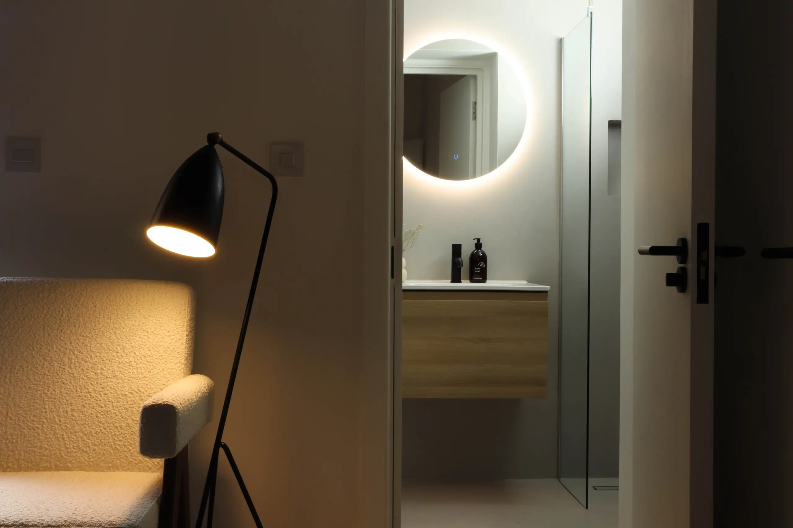 Bedroom lit by a black floor lamp beside a cream bouclé armchair, with the door open to an en-suite with a backlit round mirror and an oak vanity