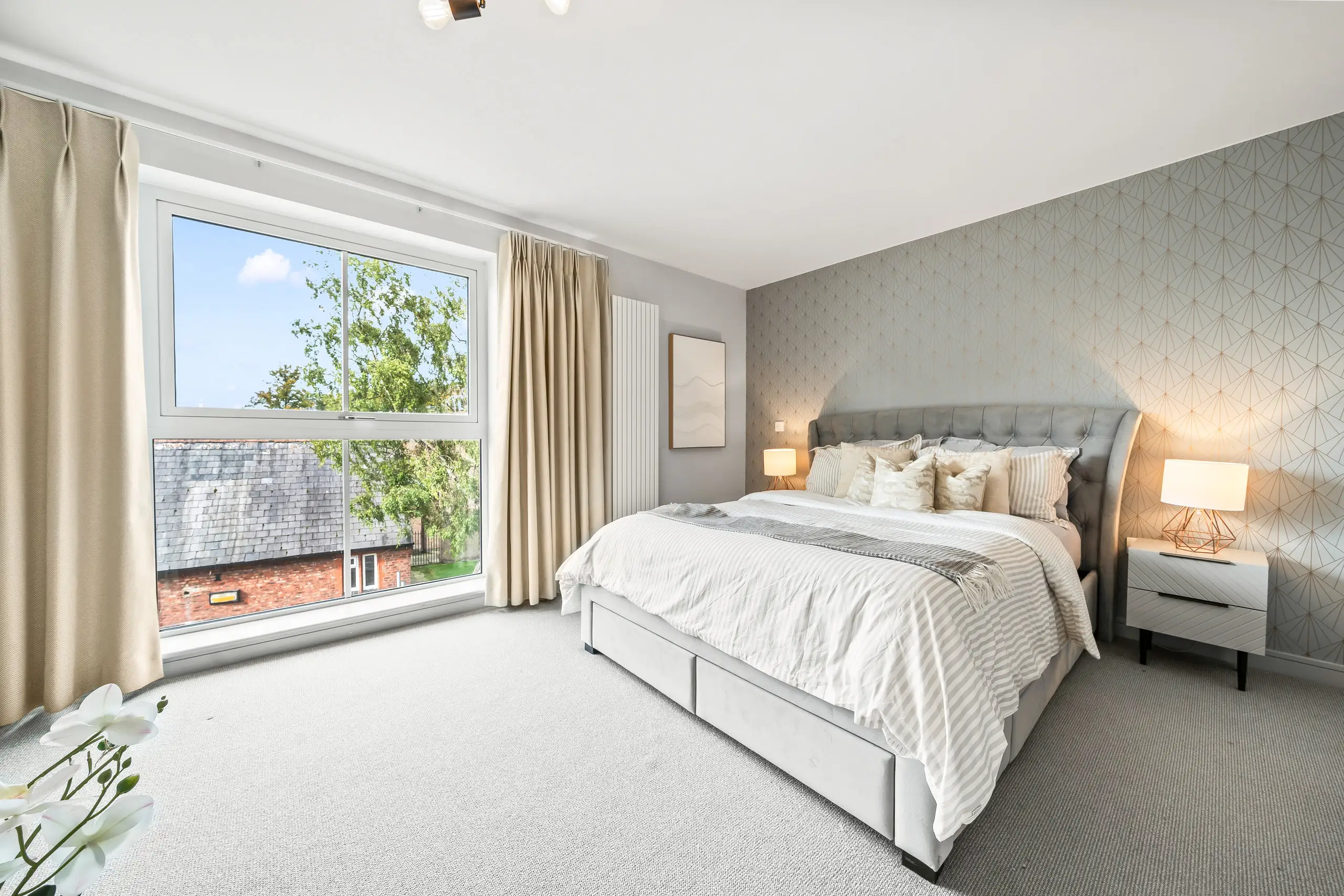 Bright bedroom with a large window, grey upholstered bed, geometric wallpaper and lit bedside lamps