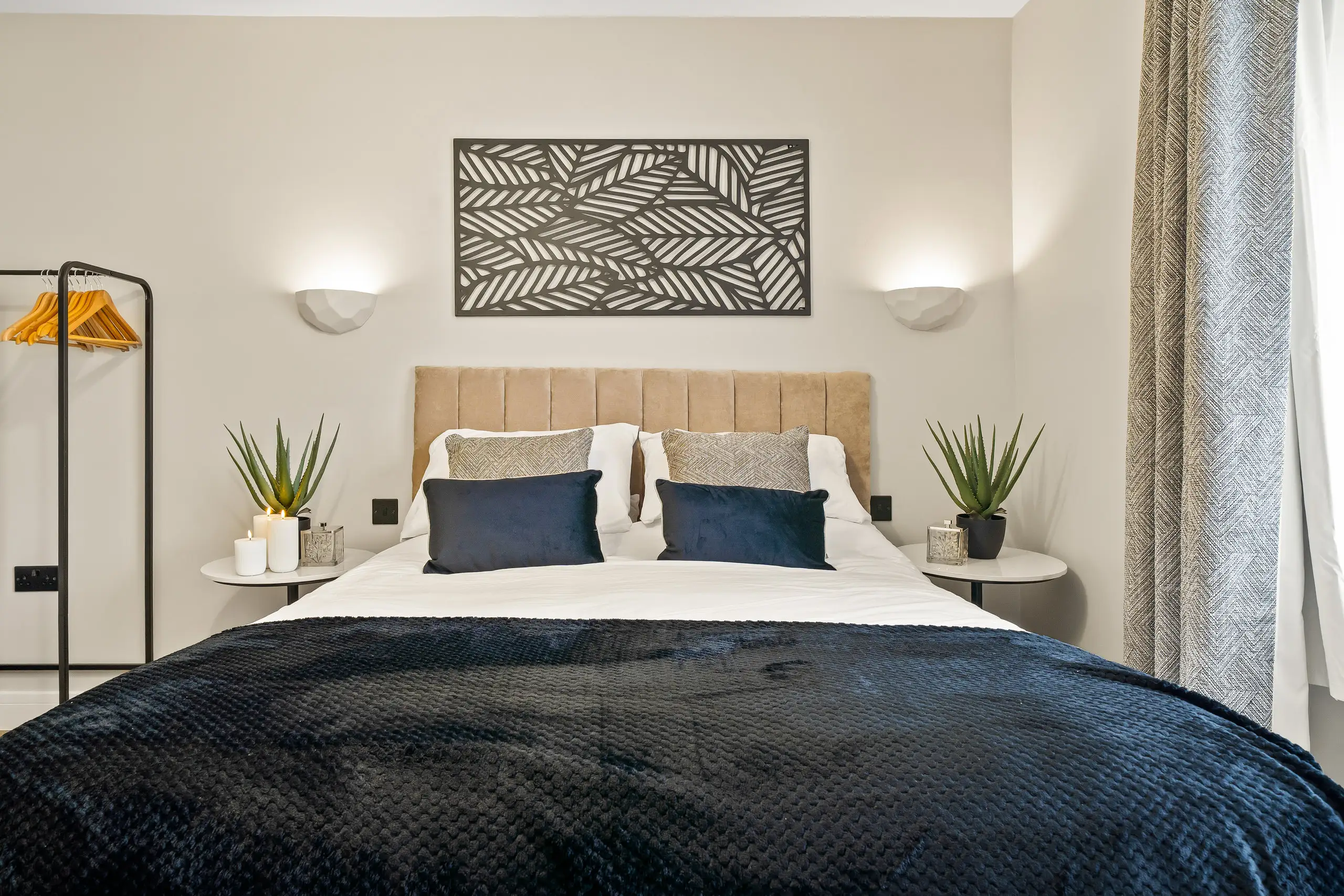 Double bedroom with a navy throw, tan headboard, wall lights and plants on matching side tables