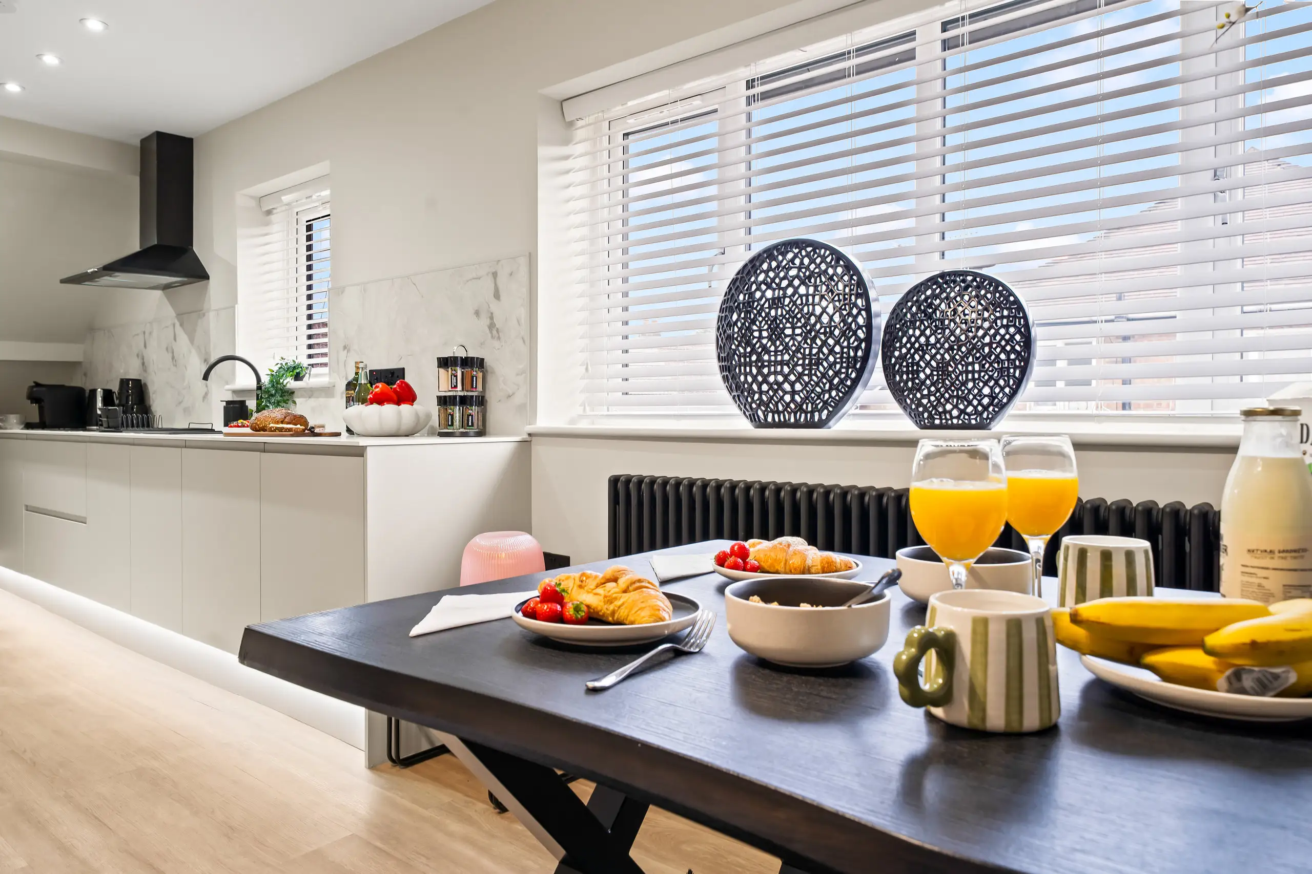 Breakfast laid on a dark dining table with orange juice, pastries and fruit, kitchen in the background