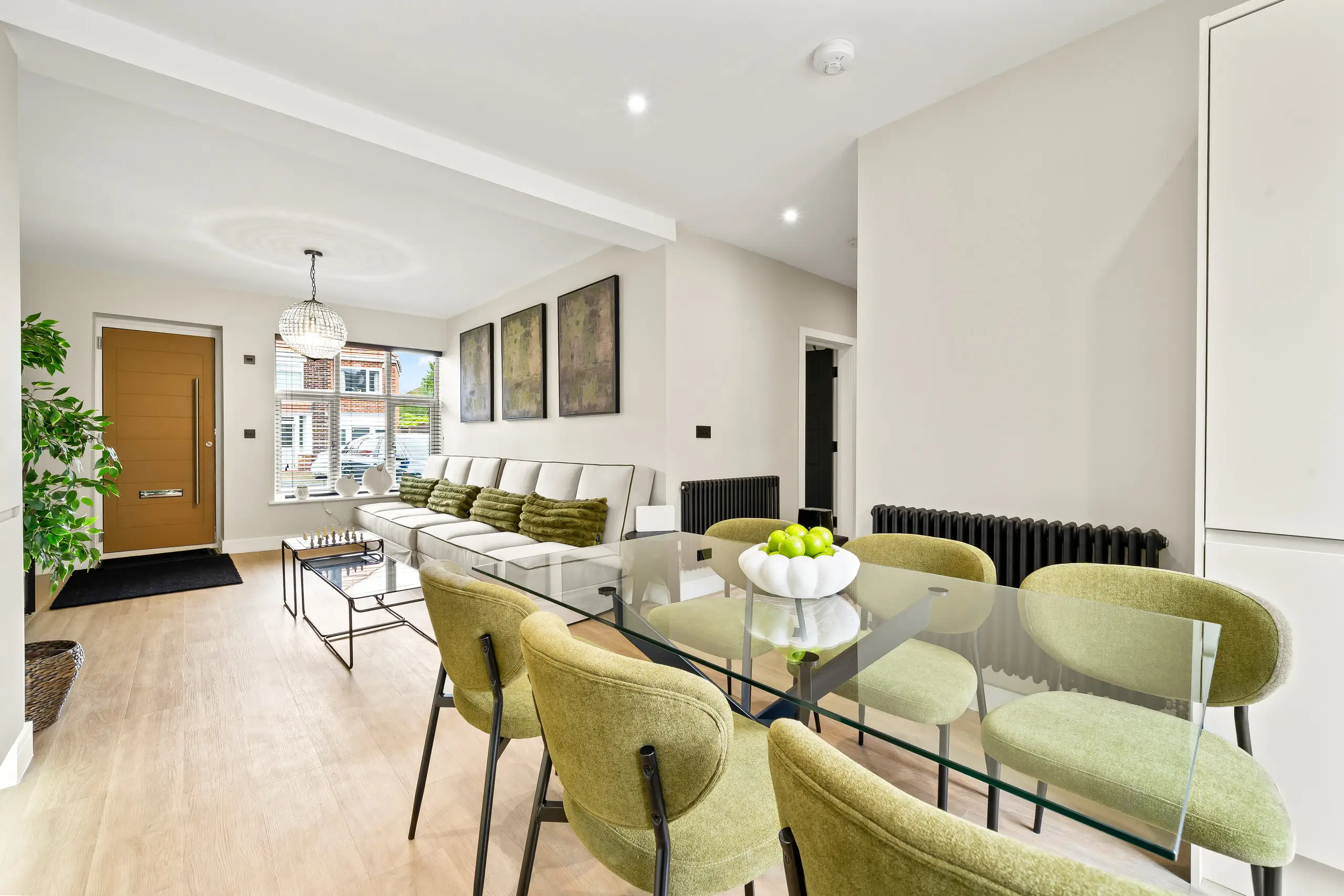 Glass dining table with green upholstered chairs in an open-plan room, with the sofa and front door beyond