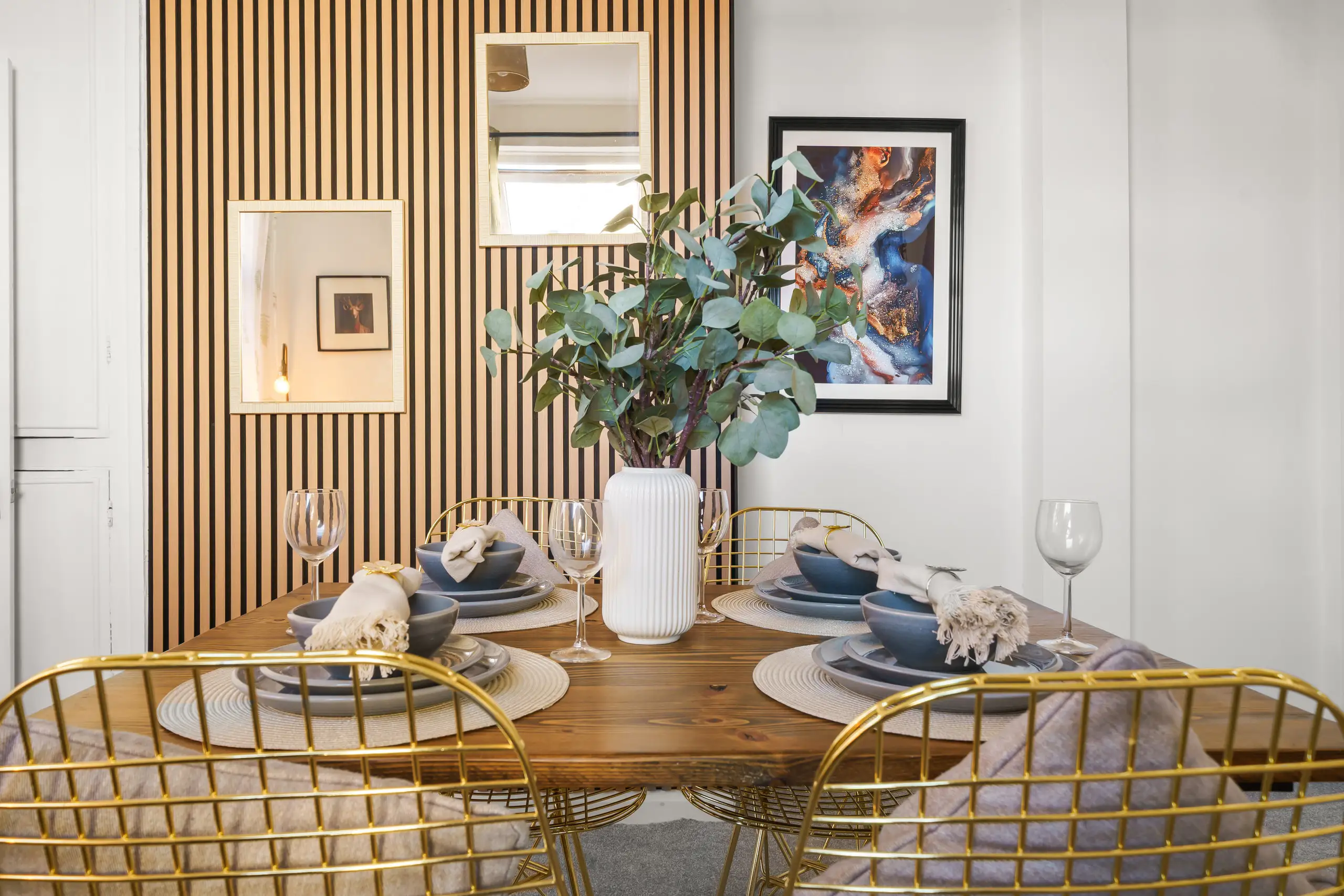 Dining table laid for four with gold wire chairs, a eucalyptus centrepiece and a slatted timber wall