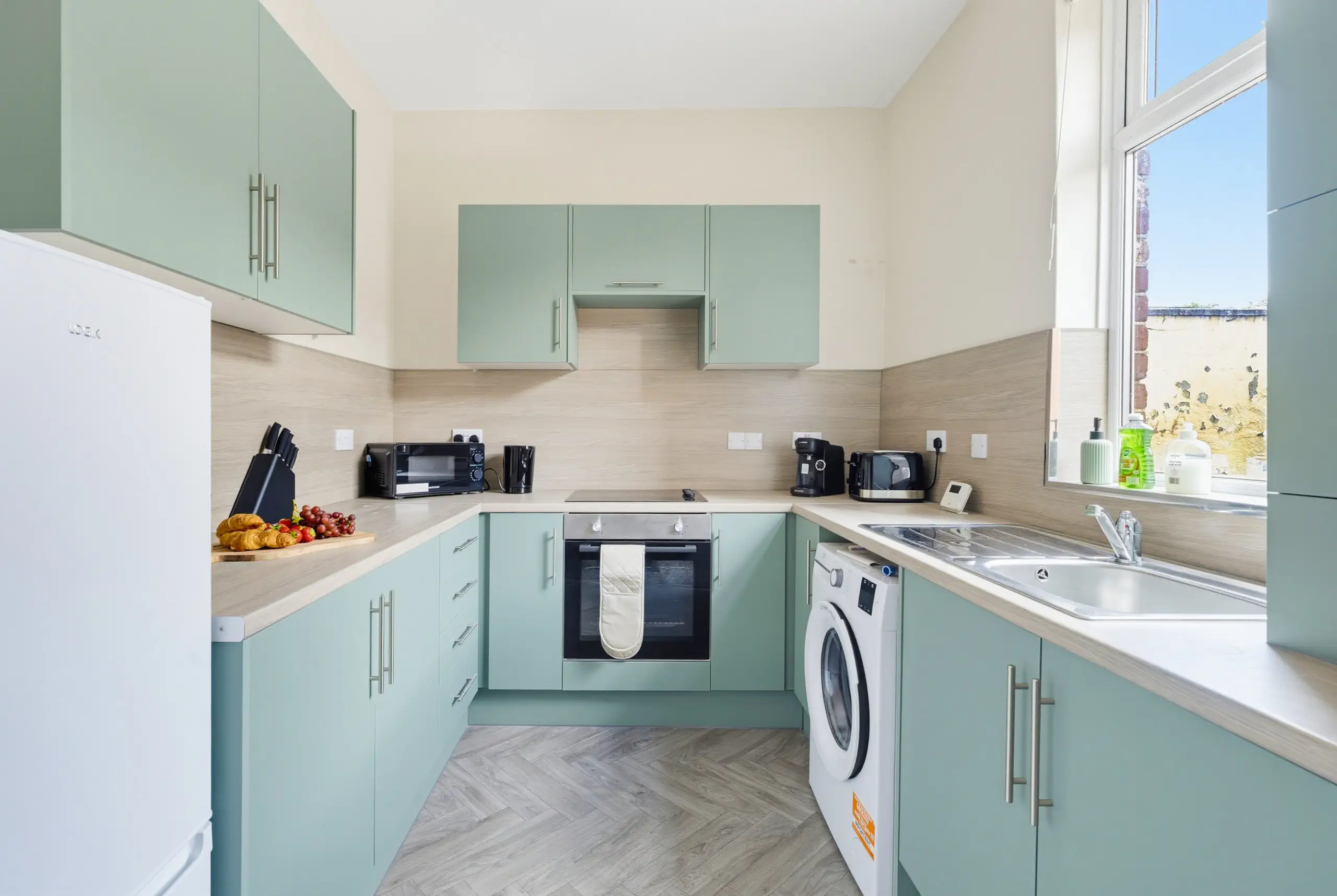 Sage green kitchen with wood-effect worktops, herringbone floor and a window above the sink