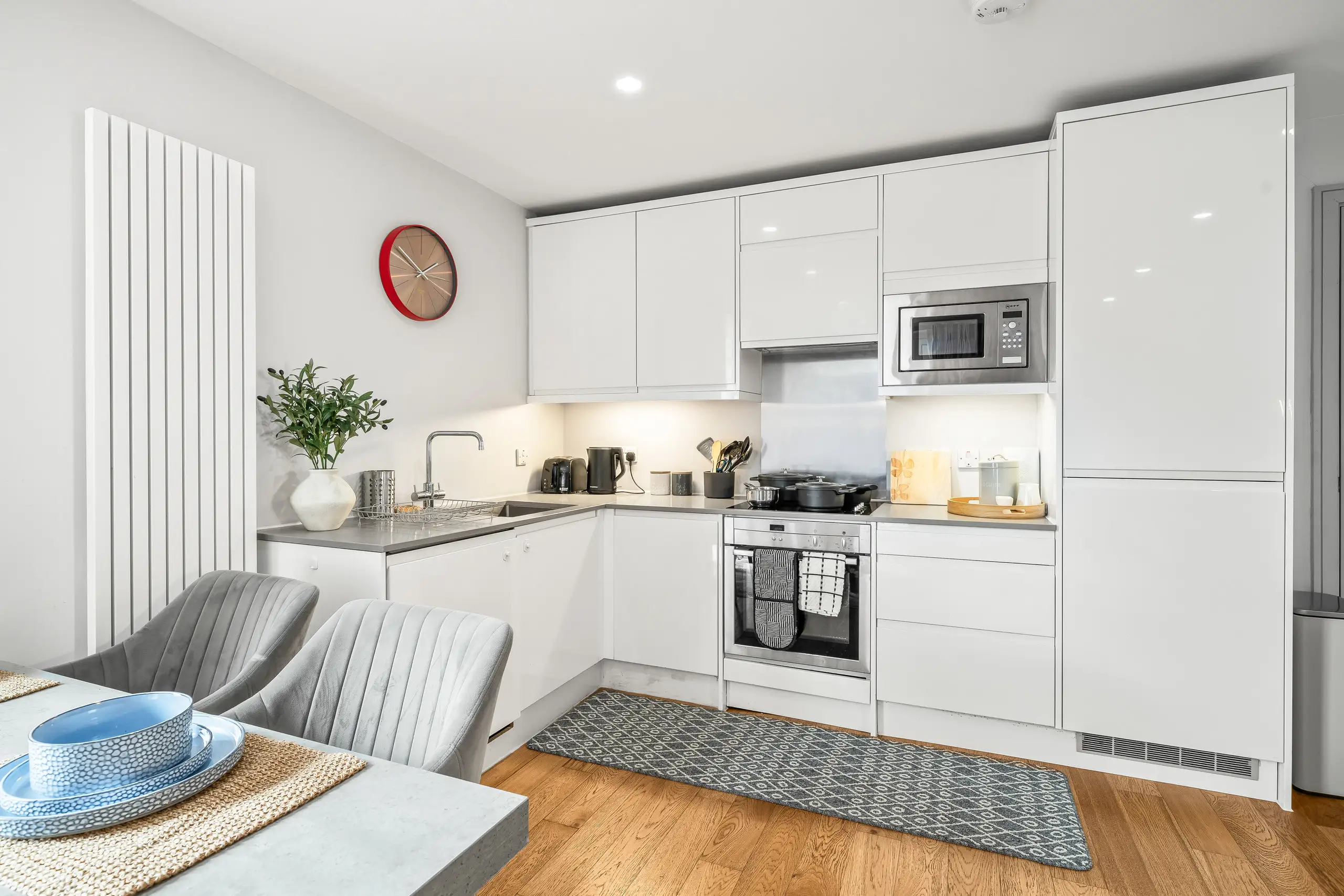 White gloss kitchen with an oak floor, grey dining chairs and a red wall clock