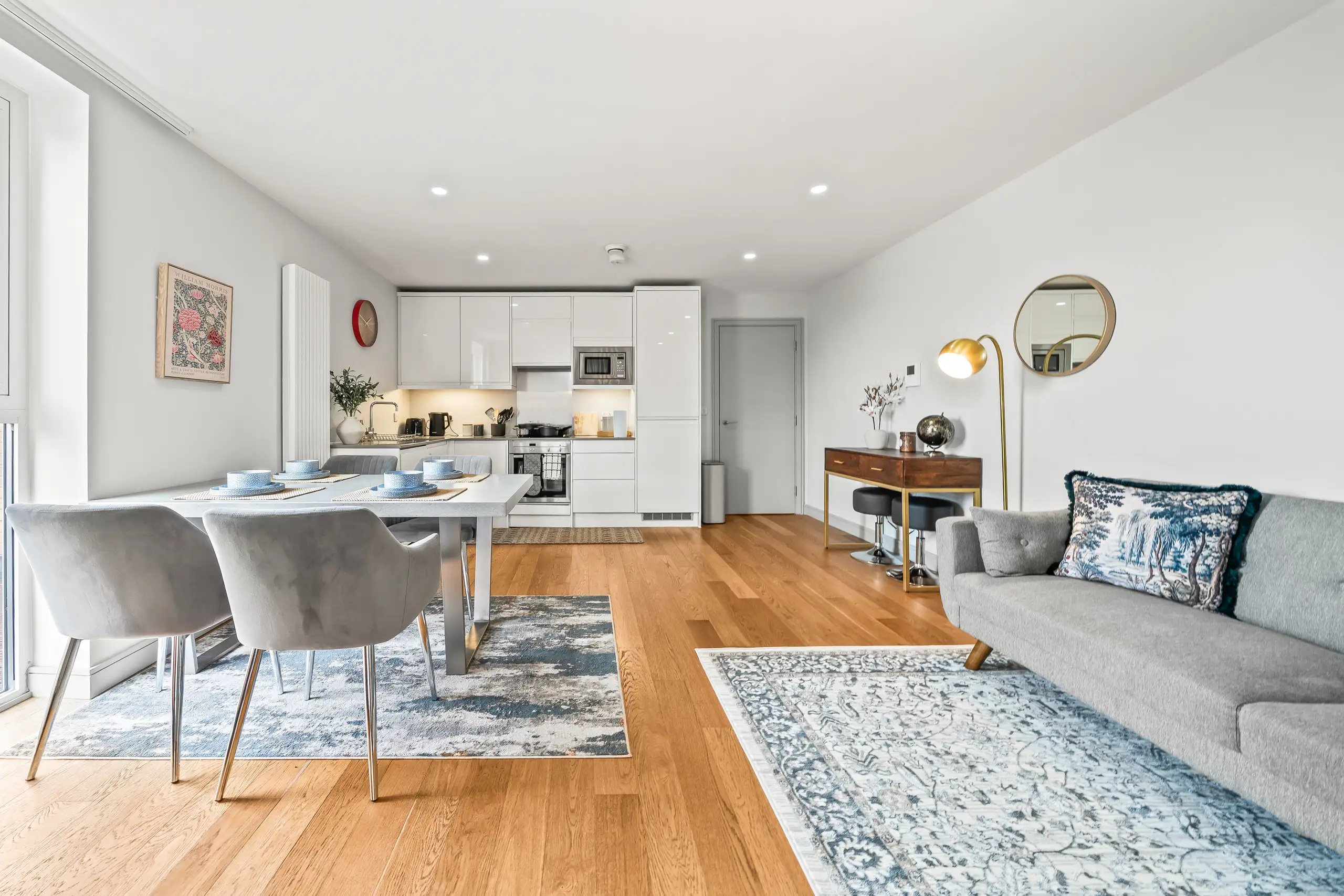 Apartment living space with an oak floor, grey sofa, patterned rugs, dining table and white kitchen at the far end