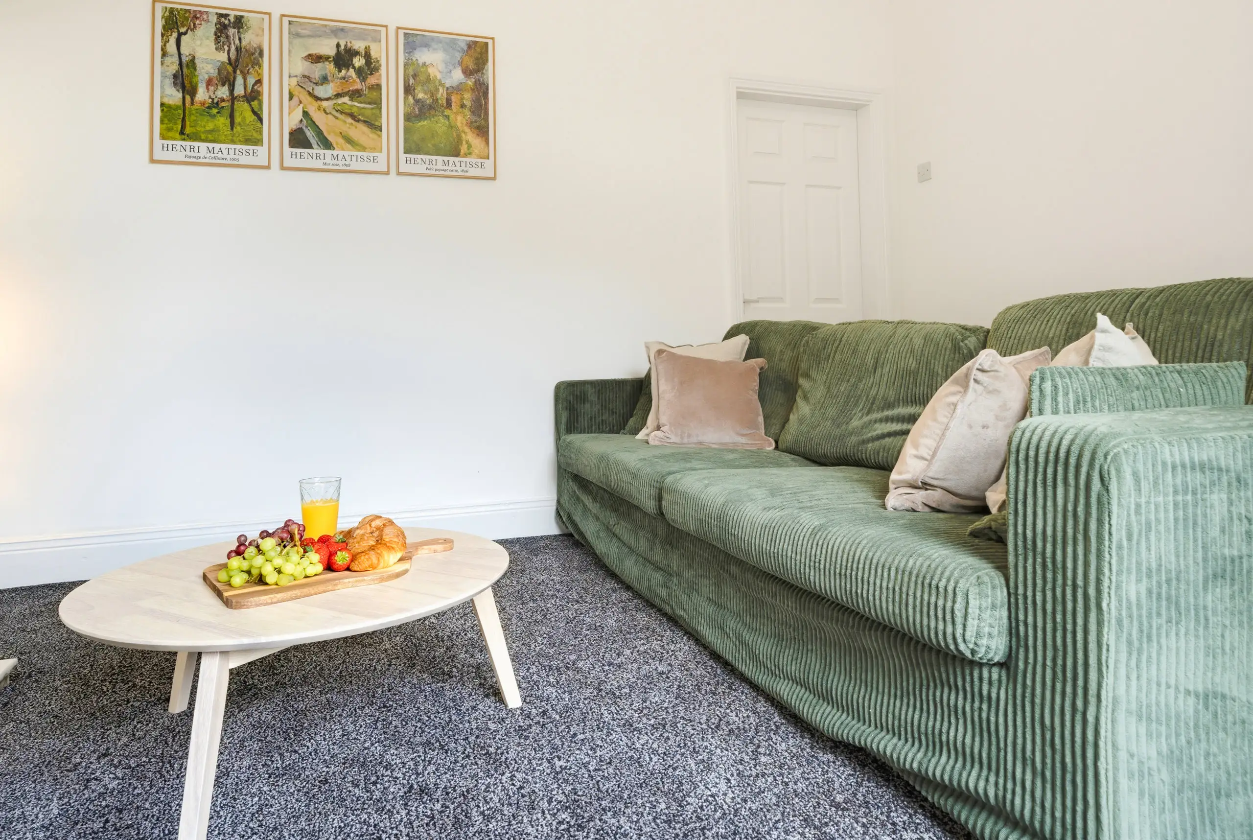 Green corduroy sofa beside a round coffee table set with fruit and pastries, three framed prints on the wall above