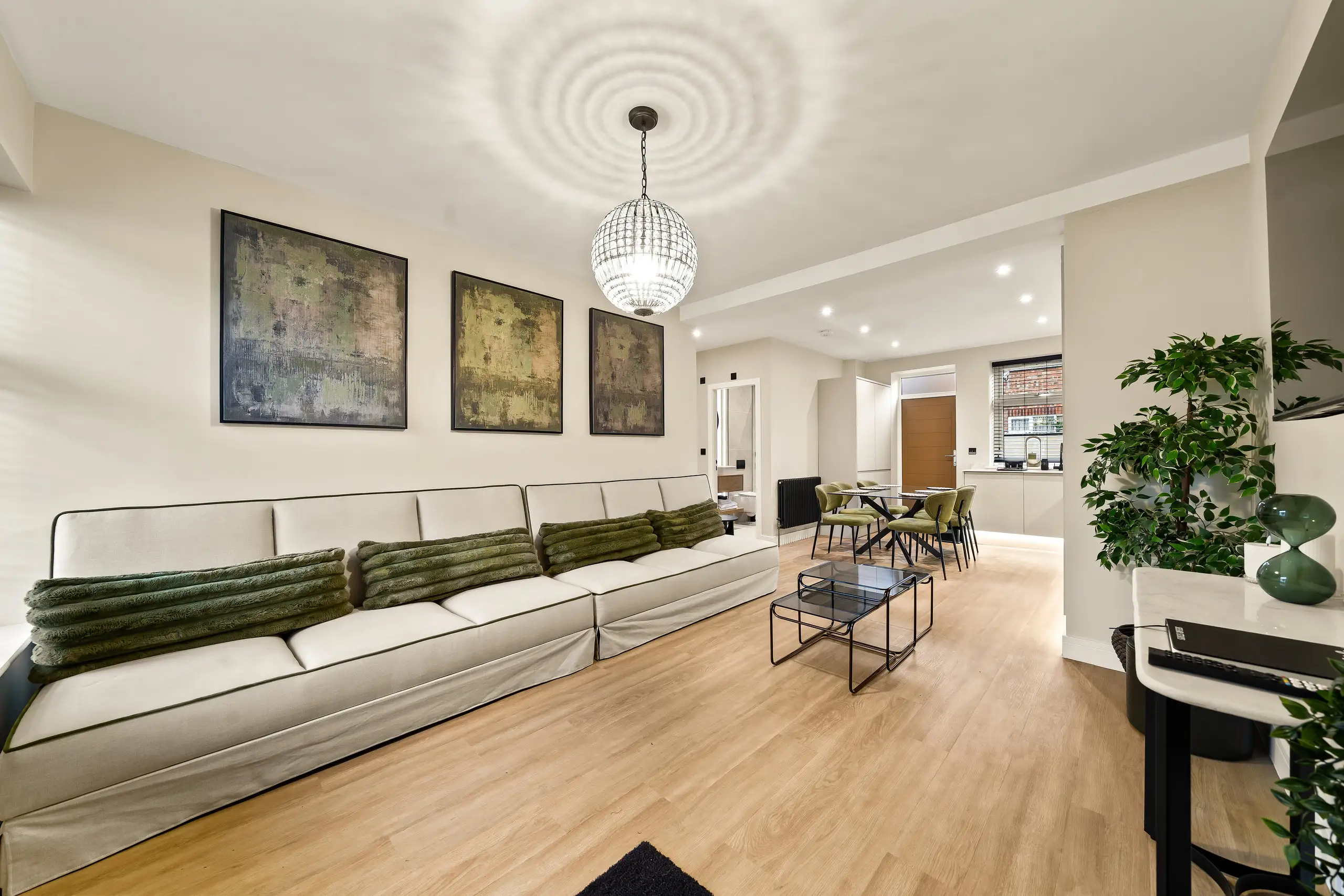 Open-plan living room with a long cream sofa, green cushions and a crystal pendant light, leading through to the dining area and kitchen