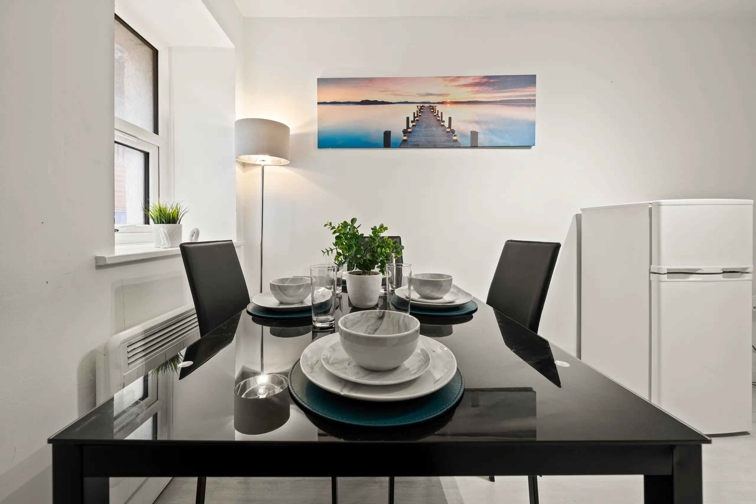 Black glass dining table laid for two, with a floor lamp and fridge freezer behind