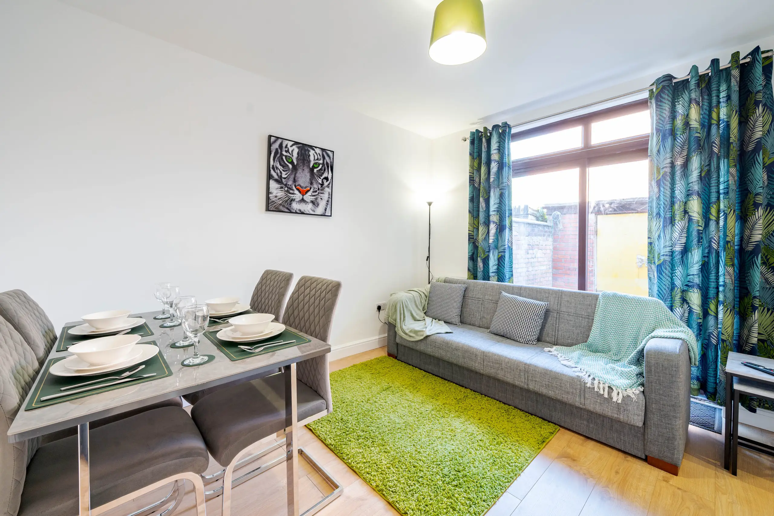 Living and dining room with a grey sofa, green rug, palm-print curtains and a table laid for four