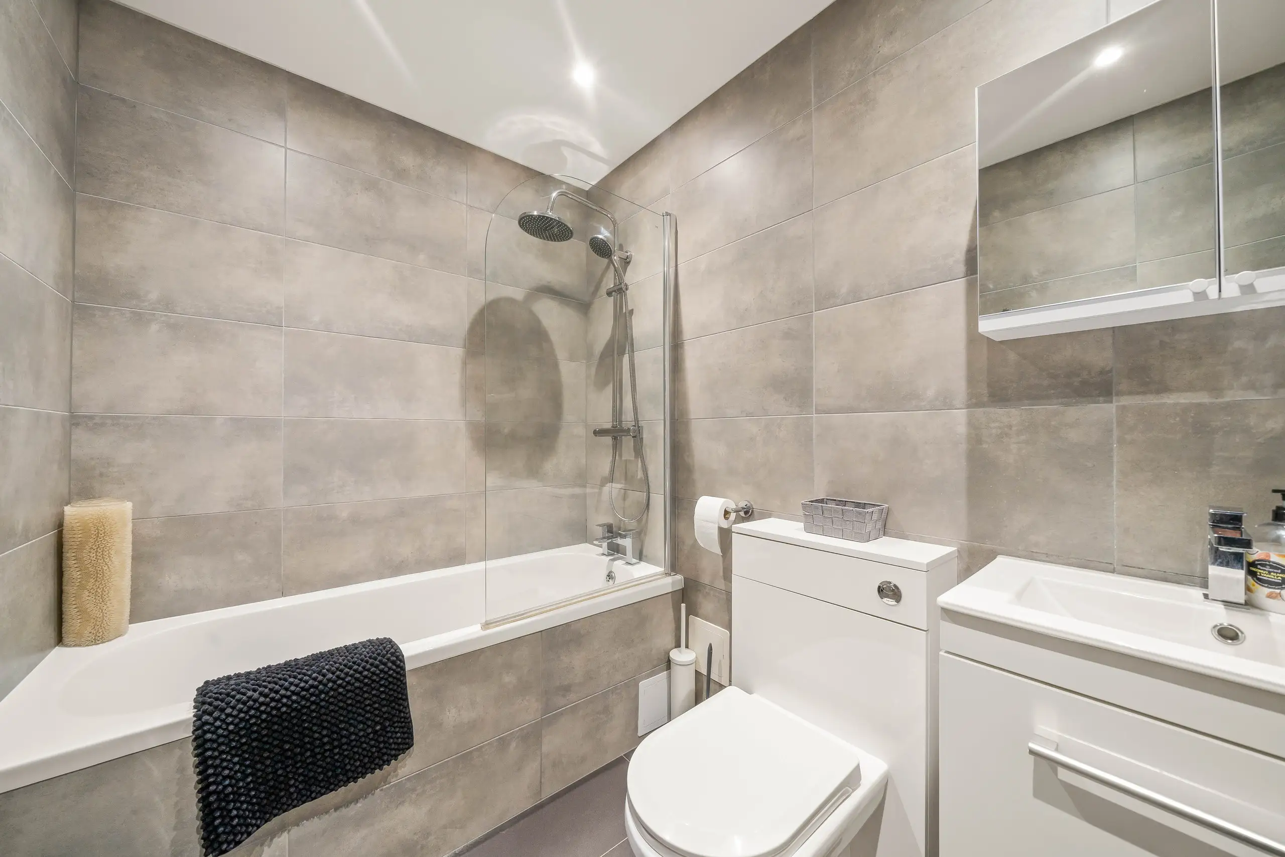 Grey-tiled bathroom with a bath and shower screen, toilet and vanity unit