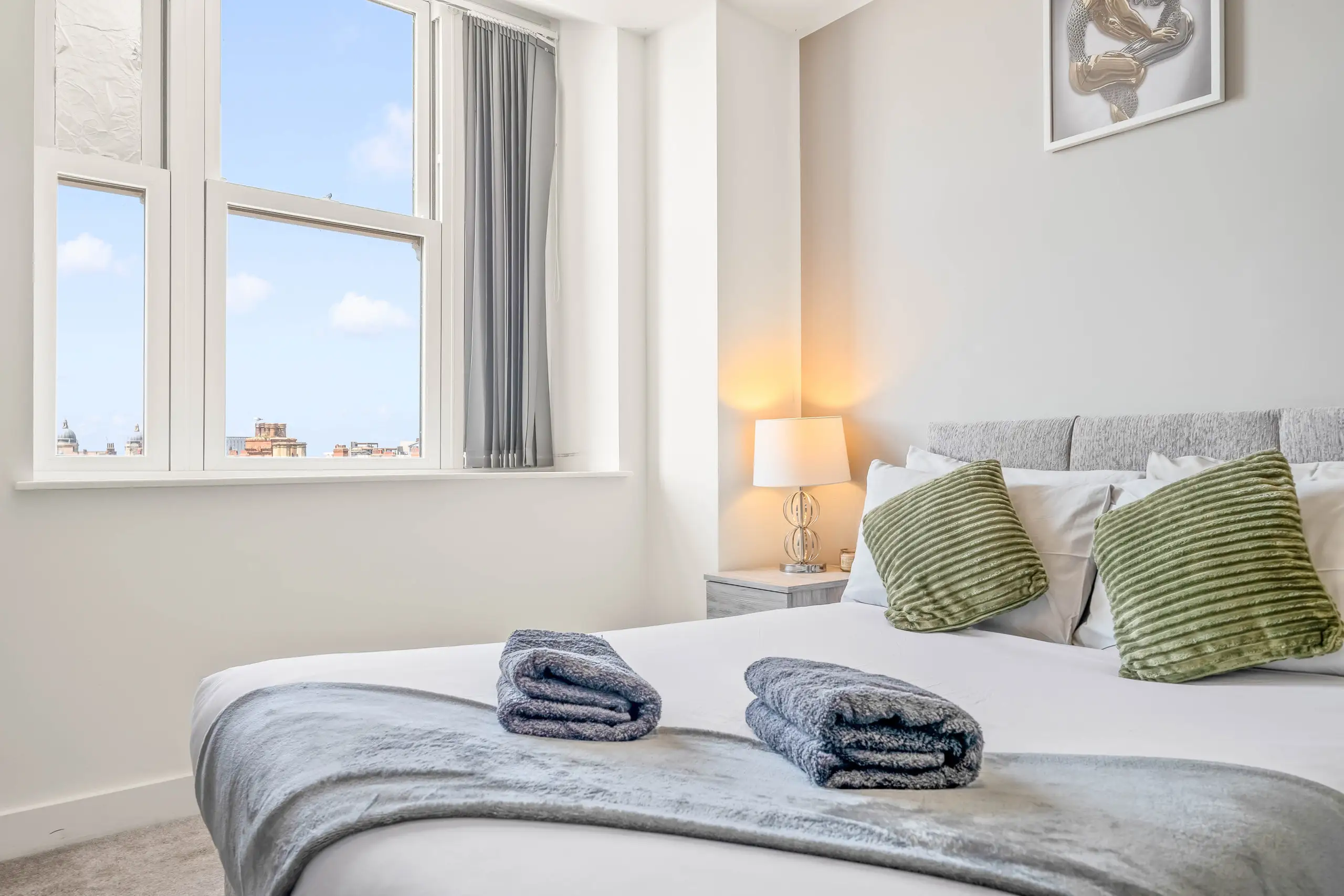 Made bed with folded towels and green cushions beside a tall window