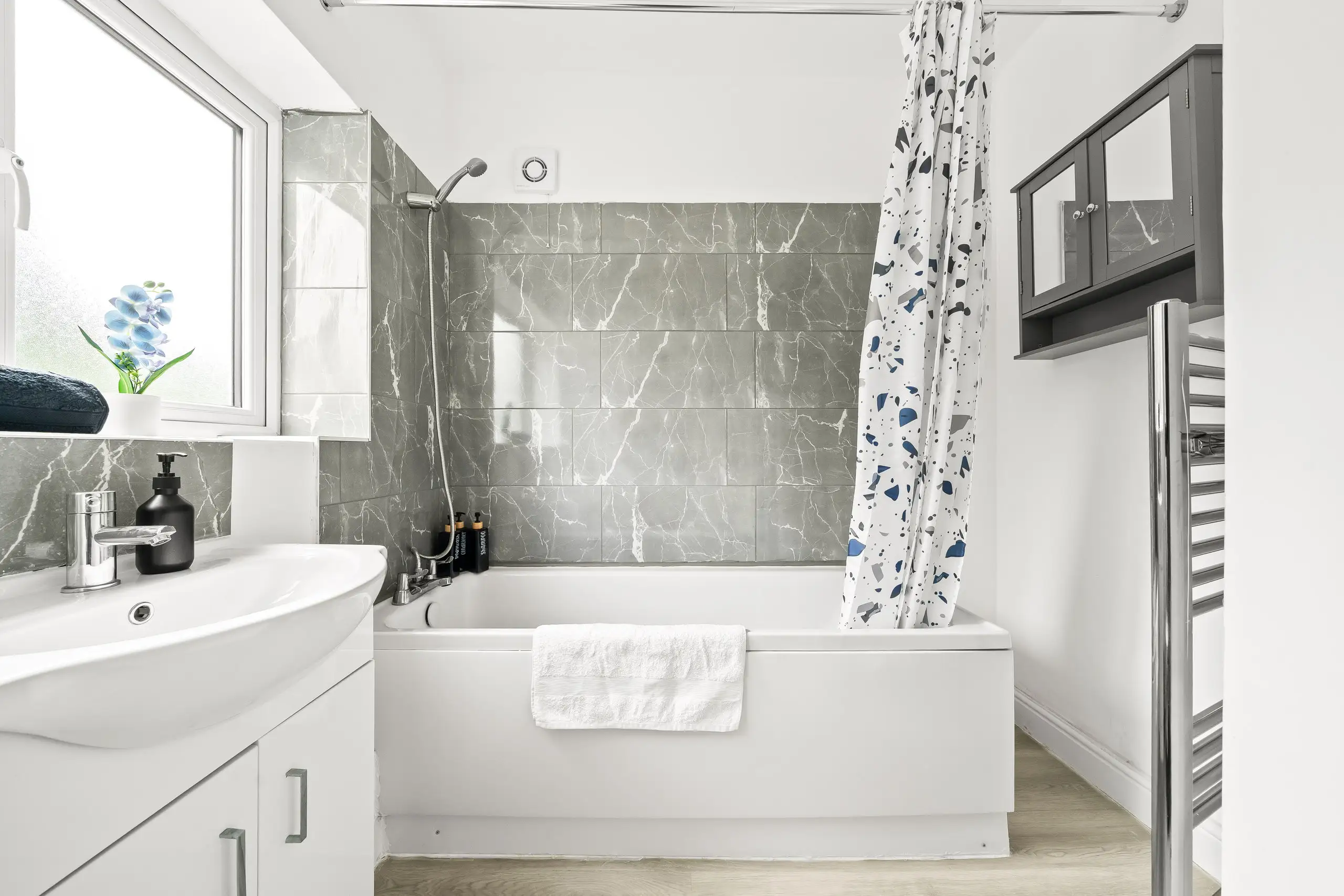 Bathroom with grey marble-effect tiles, a bath with shower, white vanity unit and towel rail
