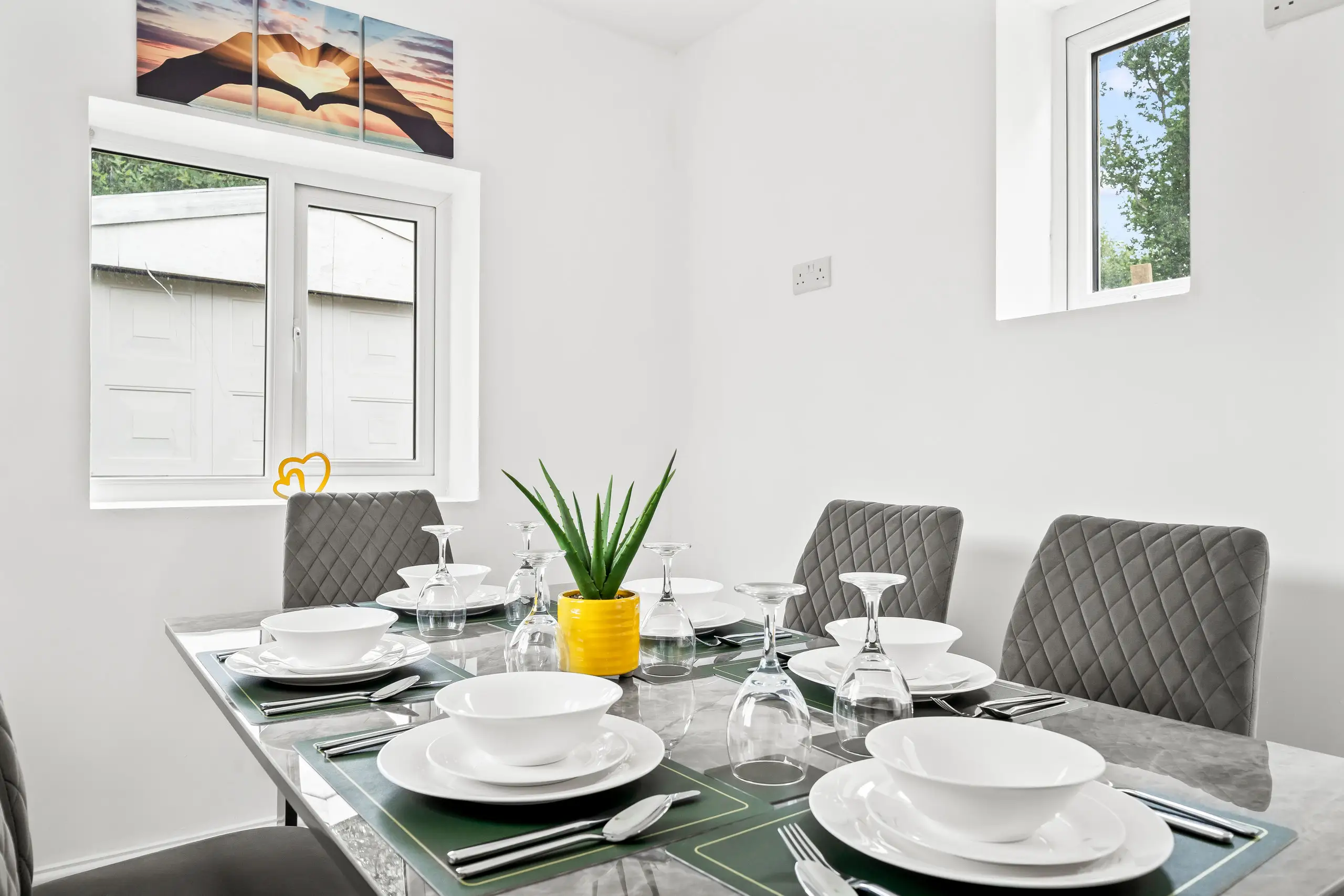 Glass dining table laid for six with grey quilted chairs and an aloe in a yellow pot