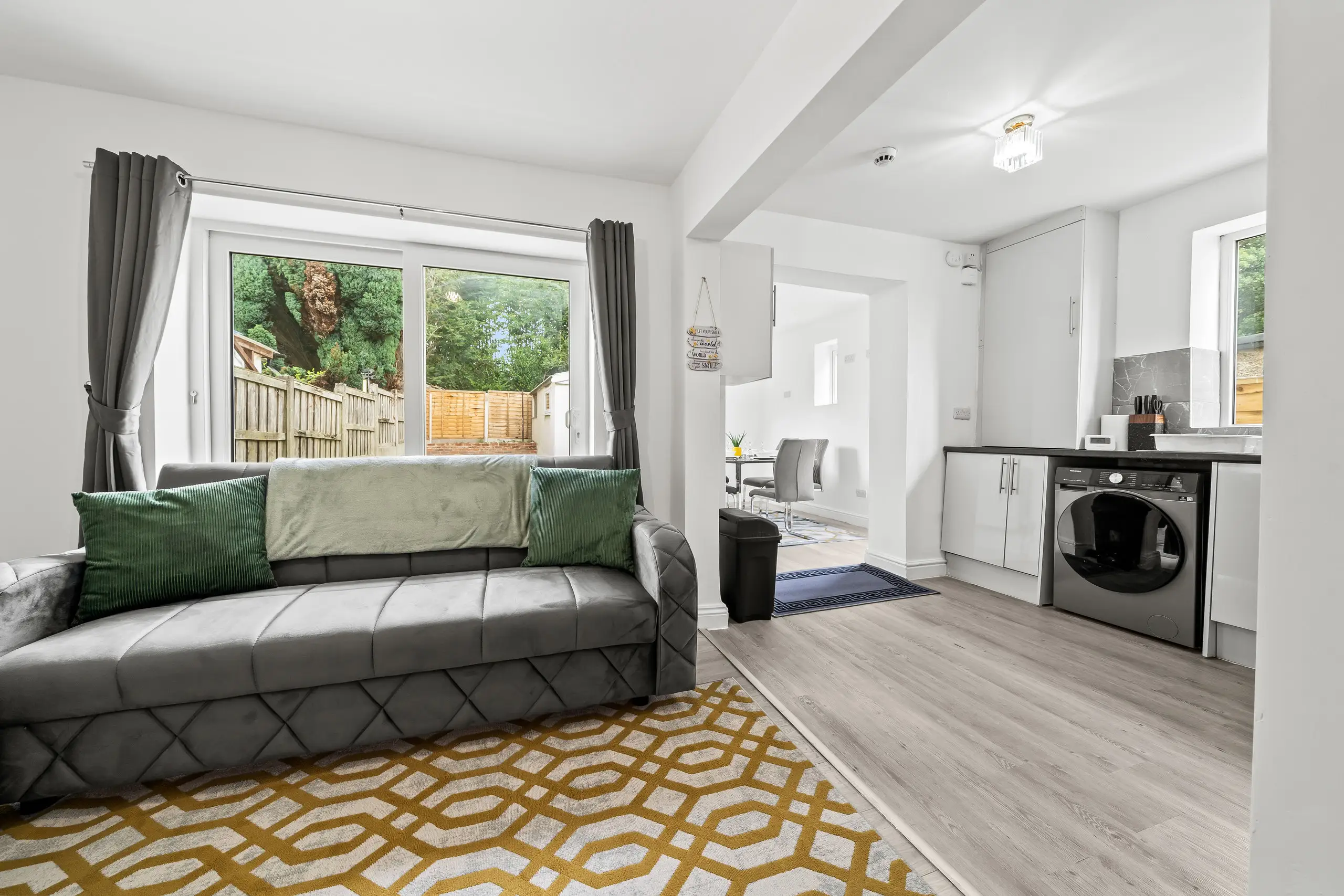 Living room with a grey sofa, green cushions and a yellow patterned rug, opening onto the kitchen