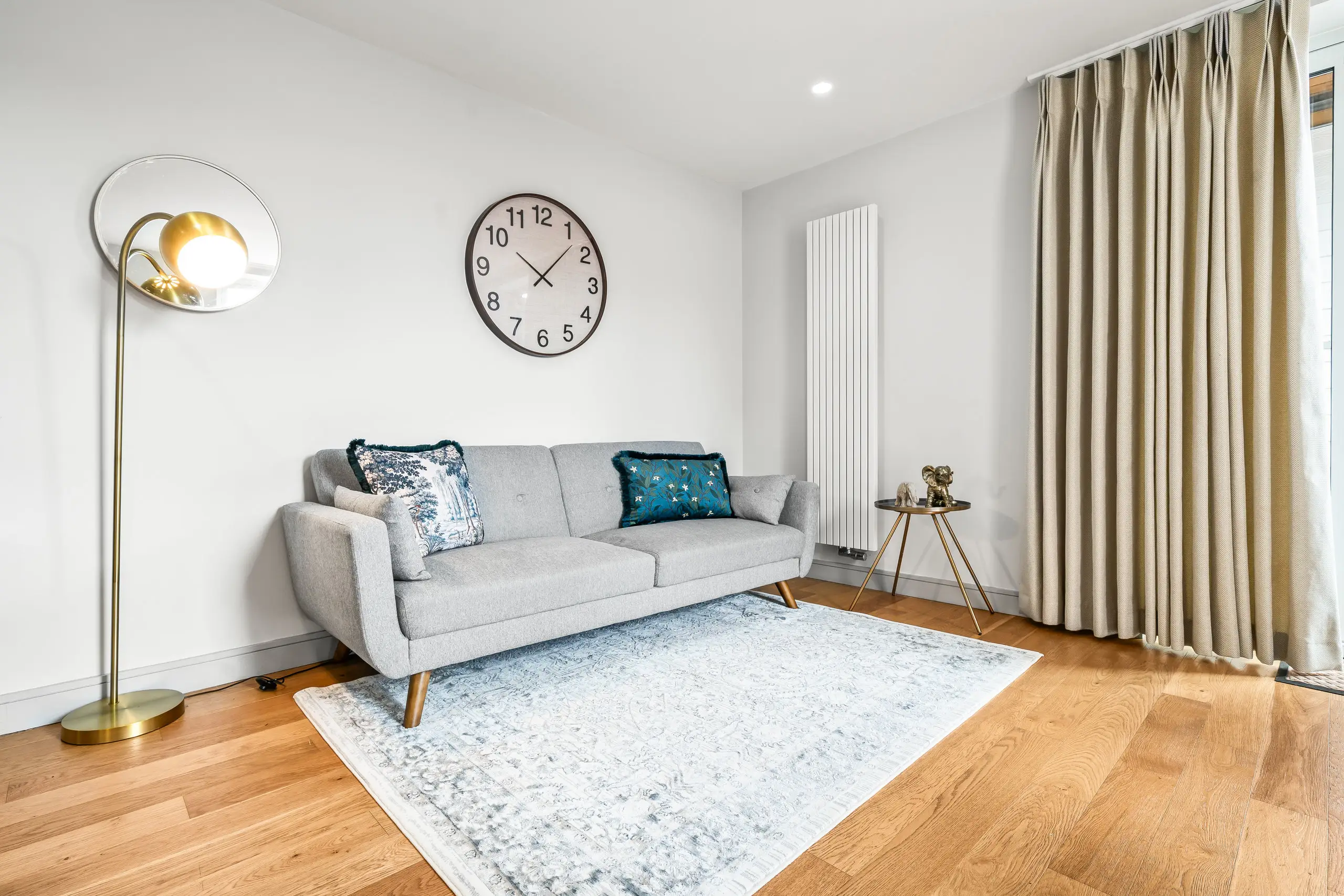 Grey sofa on an oak floor with a brass floor lamp, wall clock and pale patterned rug