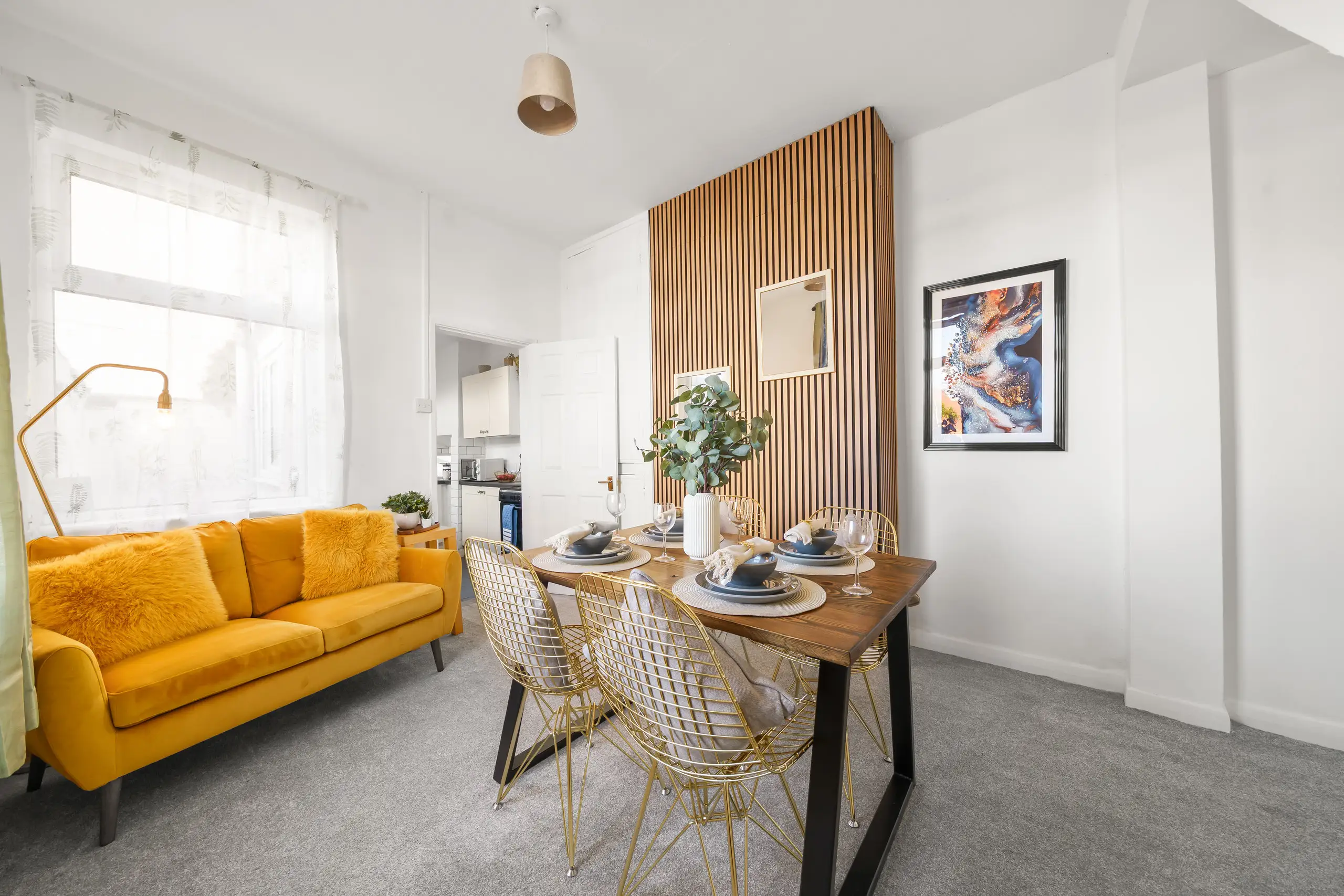 Living and dining room with a mustard velvet sofa, gold wire chairs and a slatted timber wall